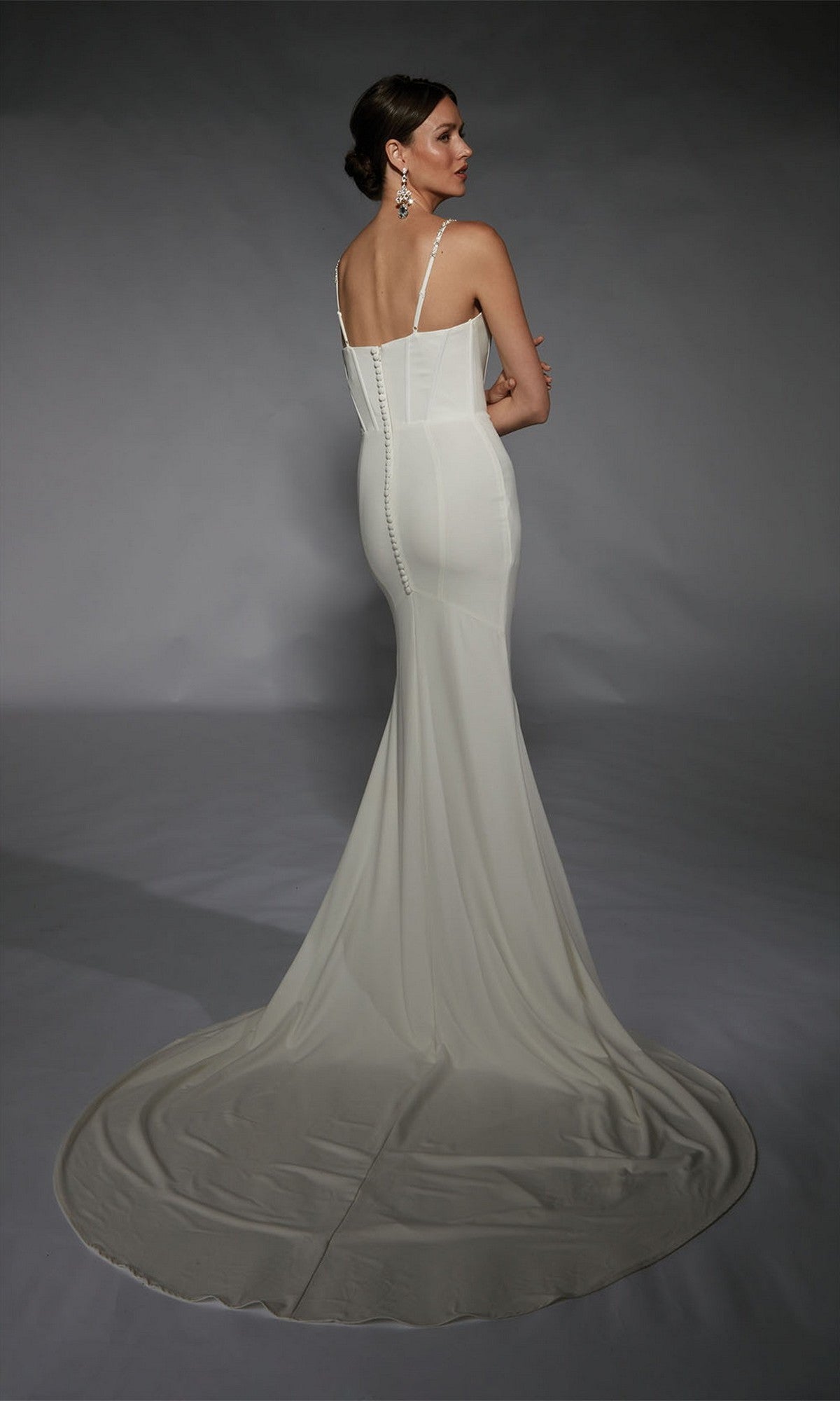 A woman stands facing away against a dark background, wearing the Long White Dress: Alyce 7104—a fitted formal gown with a long train.